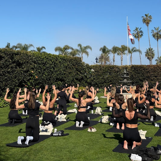 A group of people participating in a mat Pilates class outdoors, with their arms raised in unison.